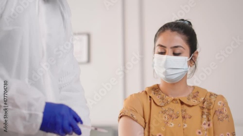 Doctor injecting a vaccination in arm of a young Indian Asian female while both faces are covered with protective masks at modern clinic or hospital amid coronavirus or COVID 19 epidemic or Pandemic