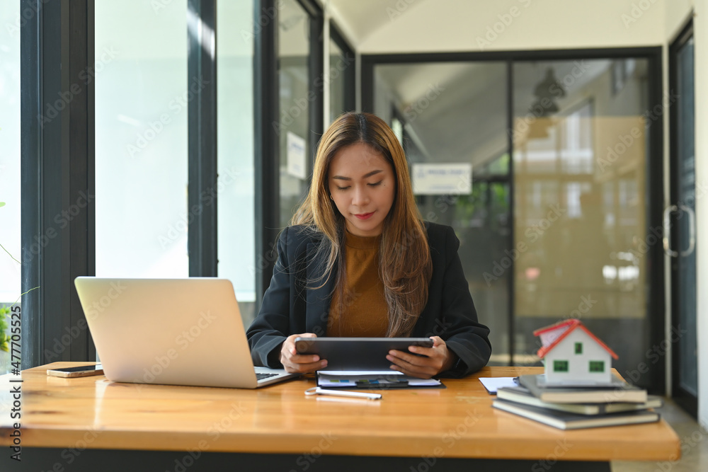 Photo of a young businesswoman holding a digital tablet while sitting at the wooden working desk surrounded by a computer laptop, house model and various office equipment.