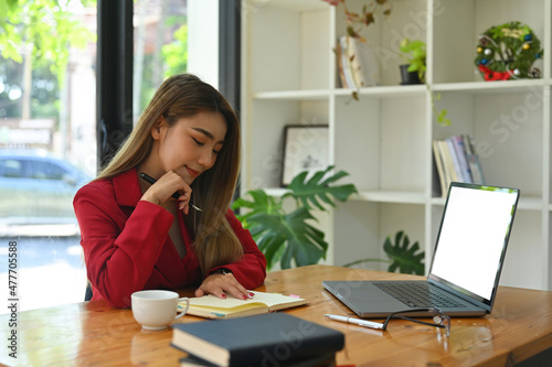 Photo of attractive woman staring at notebook on the wooden table surrounded by a white blank screen computer laptop, pen, coffee cup, eyeglasses and stack of books. © saltdium
