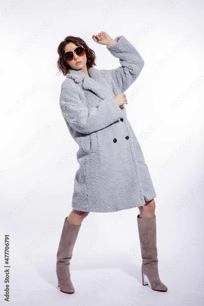 High fashion photo of a beautiful elegant young woman in a pretty pale blue fur coat, suede boots, stylish sunglasses posing over white background. Make up, hairstyle. Slim figure. High fashion photo of a beautiful elegant young woman in a pretty pale blue fur coat, suede boots, stylish sunglasses posing over white background. Make up, hairstyle. Slim figure.