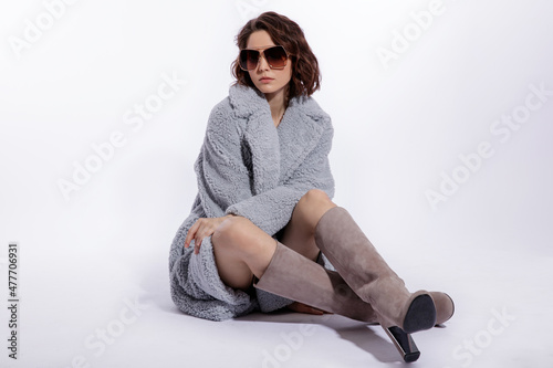 High fashion photo of a beautiful elegant young woman in a pretty pale blue fur coat, suede boots, stylish sunglasses posing over white background. Make up, hairstyle. Slim figure. Model is sitting