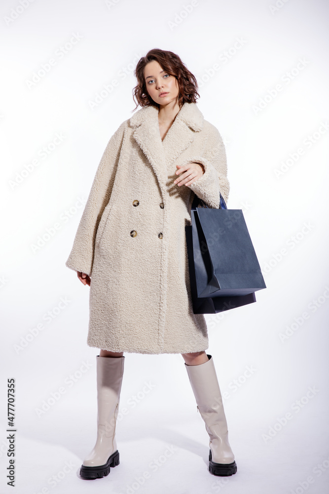 High fashion photo of a beautiful elegant young woman in a pretty cream beige fur coat, boots, two blue shopping bags posing over white background. Make up, hairstyle. Slim figure. High fashion photo of a beautiful elegant young woman in a pretty cream beige fur coat, boots, two blue shopping bags posing over white background. Make up, hairstyle. Slim figure.