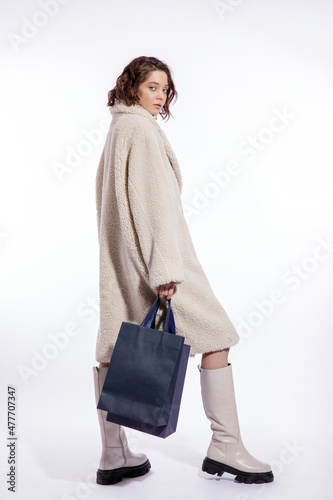 High fashion photo of a beautiful elegant young woman in a pretty cream beige fur coat, boots, two blue shopping bags posing over white background. Make up, hairstyle. Slim figure.