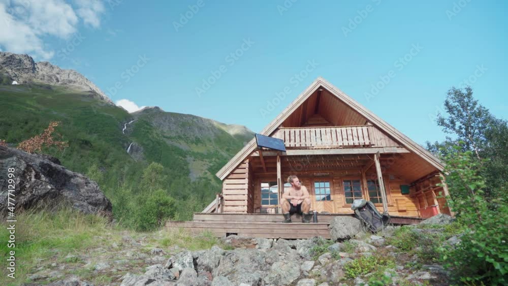 Shirtless Man Tourist Relaxing Sitting On The Front Porch Steps Of A Wooden Cabin In Lyngsdalen Valley, Norway. - Low-Angle Shot Shirtless Man Tourist Relaxing Sitting On The Front Porch Steps Of A Wooden Cabin In Lyngsdalen Valley, Norway. - Low-Angle Shot
