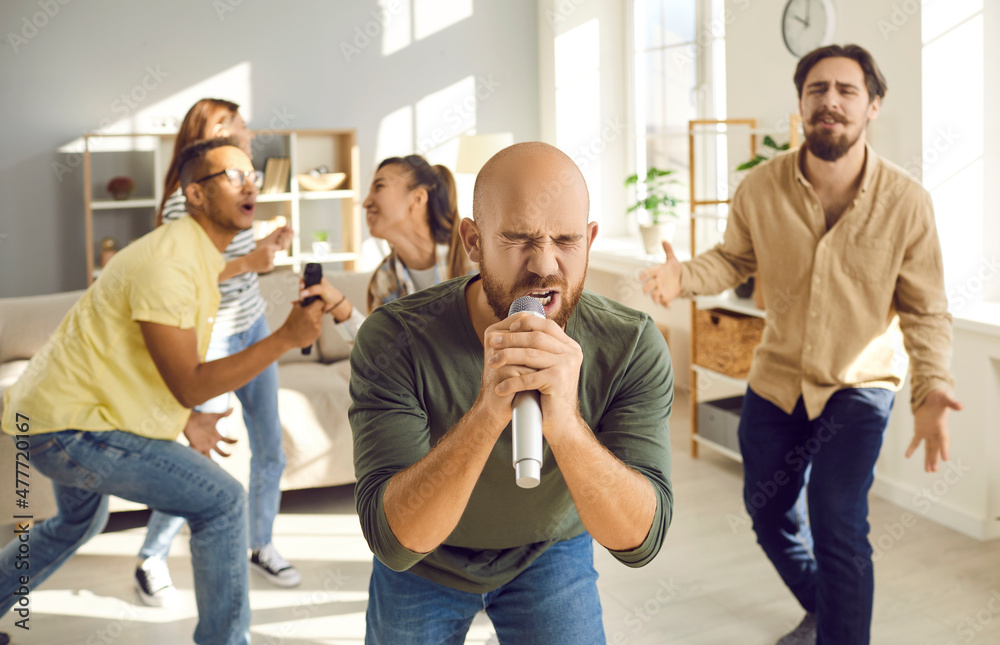 Obraz premium Karaoke at home. Emotional young man holding a microphone and enthusiastically singing while having fun with friends at home. Man sings against background of friends who also sing and have fun in room