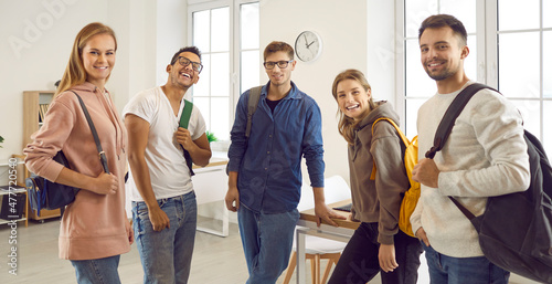 Indoor group portrait of happy positive diverse fellow students. Five confident mixed race multiethnic university friends with bags standing together in modern classroom, smiling and looking at camera