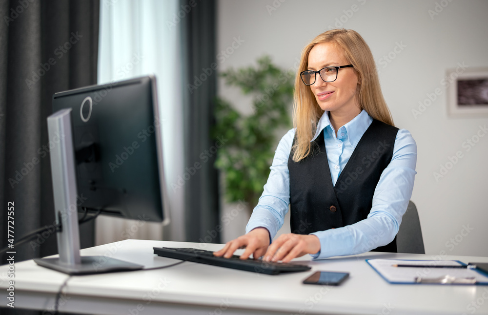 Middle aged caucasian woman in formal clothes and eyeglasses typing on modern computer while sitting at home office. Concept of freelance and technology. Middle aged caucasian woman in formal clothes and eyeglasses typing on modern computer while sitting at home office. Concept of freelance and technology.