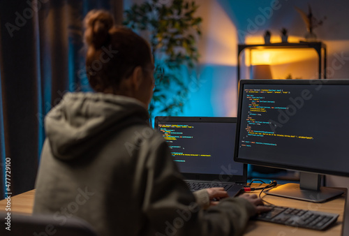 Back view of woman in casual hoodie sitting at workplace and doing programming on two modern computers. Female freelancer reading data codes during evening time at home.