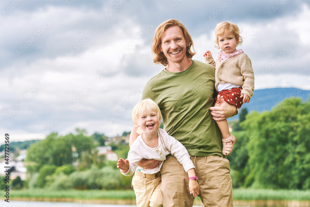 Fototapeta premium Outdoor portrait of happy young father holding two children in arms, toddler girl and preschooler boy having fun with dad on a nice sunny day