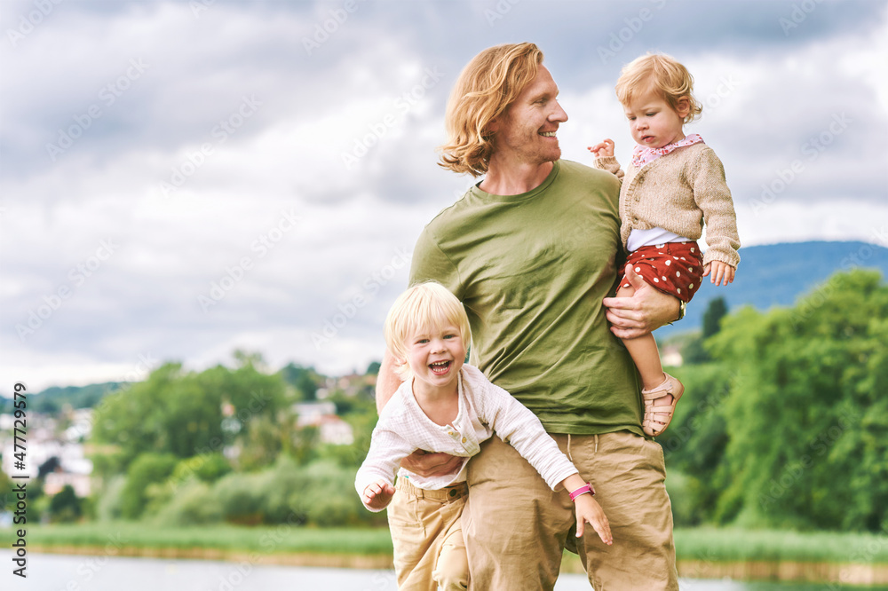 Fototapeta premium Outdoor portrait of happy young father holding two children in arms, toddler girl and preschooler boy having fun with dad on a nice sunny day