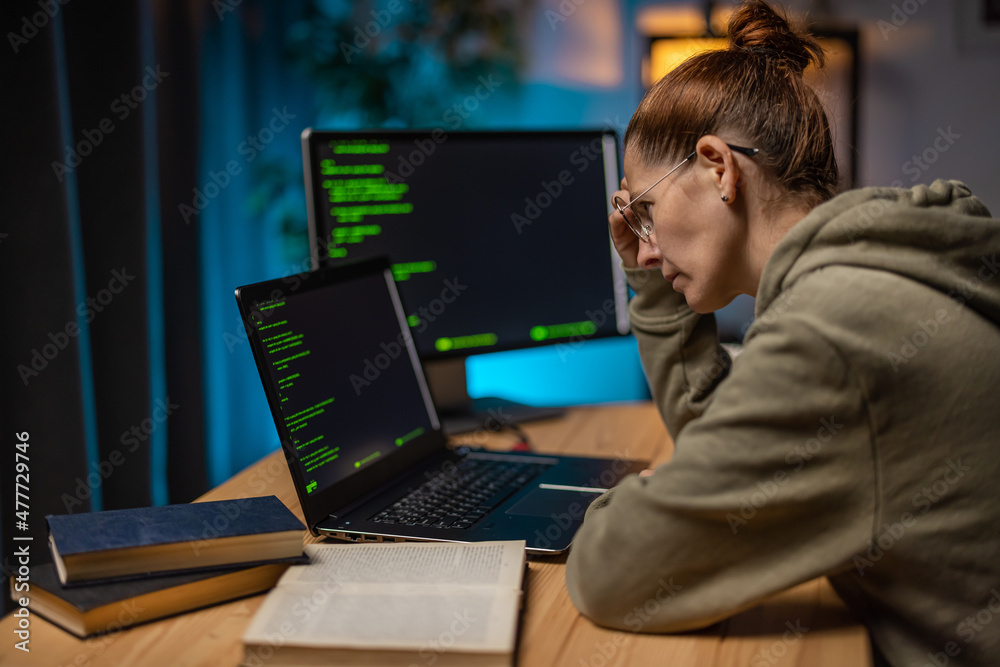 Side view of focused caucasian woman in eyewear sitting at desk with laptop and pc while examining html code on screens. Late work at home of female programmer. Side view of focused caucasian woman in eyewear sitting at desk with laptop and pc while examining html code on screens. Late work at home of female programmer.