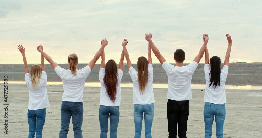 Team of friends wearing jeans and white t-shirts raises hands holding together and standing on bank of river under cloudy sky backside view Team of friends wearing jeans and white t-shirts raises hands holding together and standing on bank of river under cloudy sky backside view