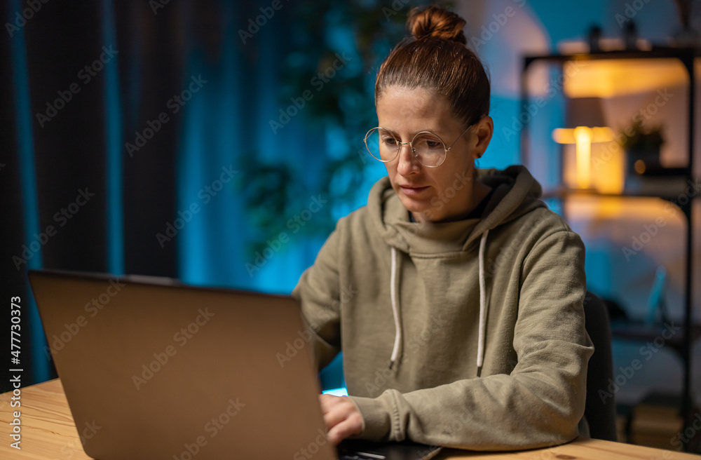 Mature woman in eyeglasses using wireless laptop while sitting at desk. Female freelance working late at home. Technology and people concept.