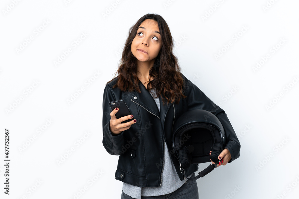 Young woman holding a motorcycle helmet over isolated white background holding coffee to take away and a mobile while thinking something Young woman holding a motorcycle helmet over isolated white background holding coffee to take away and a mobile while thinking something