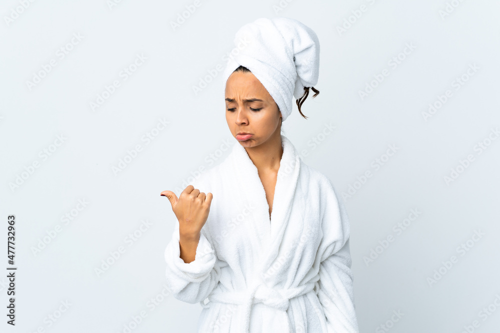 Young woman in bathrobe over isolated white background unhappy and pointing to the side Young woman in bathrobe over isolated white background unhappy and pointing to the side