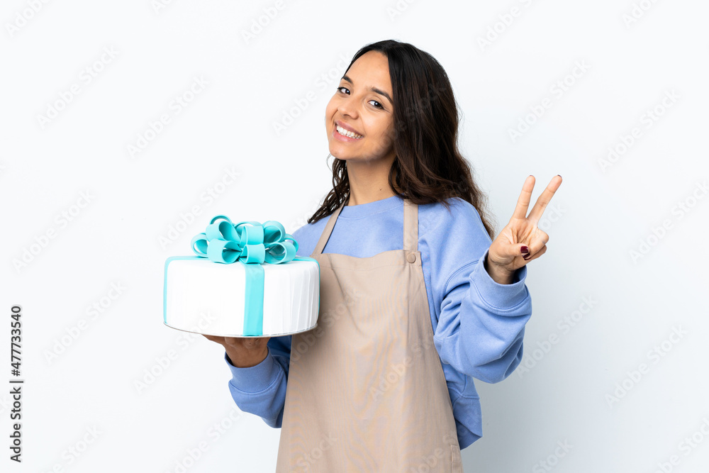 Pastry chef holding a big cake over isolated white background smiling and showing victory sign Pastry chef holding a big cake over isolated white background smiling and showing victory sign