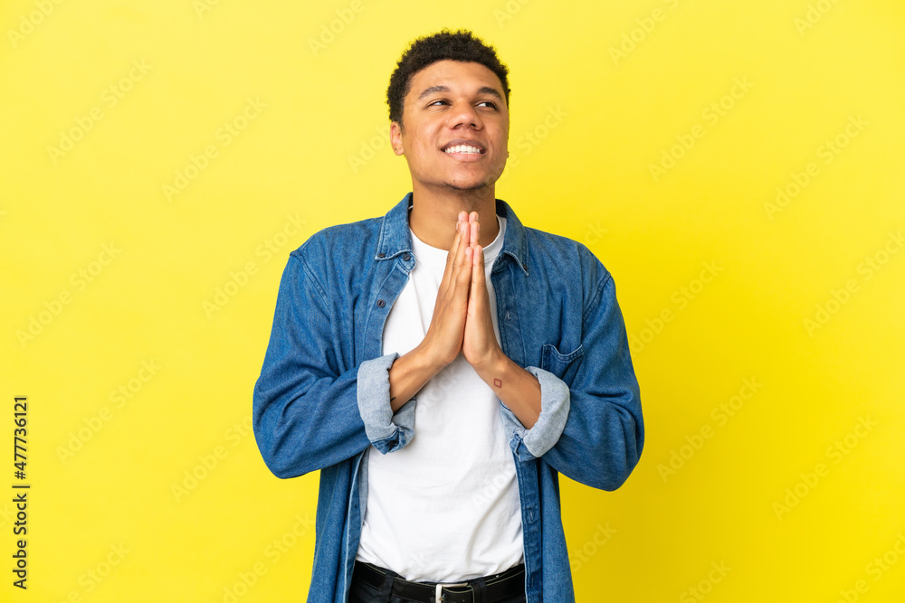 Young African American man isolated on yellow background keeps palm together. Person asks for something