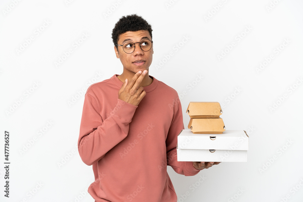 Young African American man holding a burger and pizzas isolated on white background looking up while smiling Young African American man holding a burger and pizzas isolated on white background looking up while smiling
