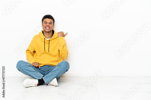 Young African American man sitting on the floor isolated on white background pointing to the side to present a product