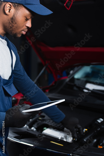 young african american repairman holding digital tablet while making inspection of blurred car.