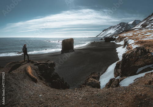 Person standing at black sand beach in Iceland. Fauskasandur near village of Djupivogur. Black sand beachwith snow and beautiful sunny weather. Seastack monolith. photo