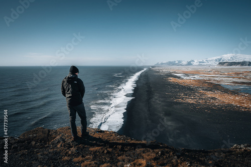 Person standing at Dyrholaey lighthouse in Iceland looking out over the black sand beach below during winter with snow and beautiful sunny weather. photo