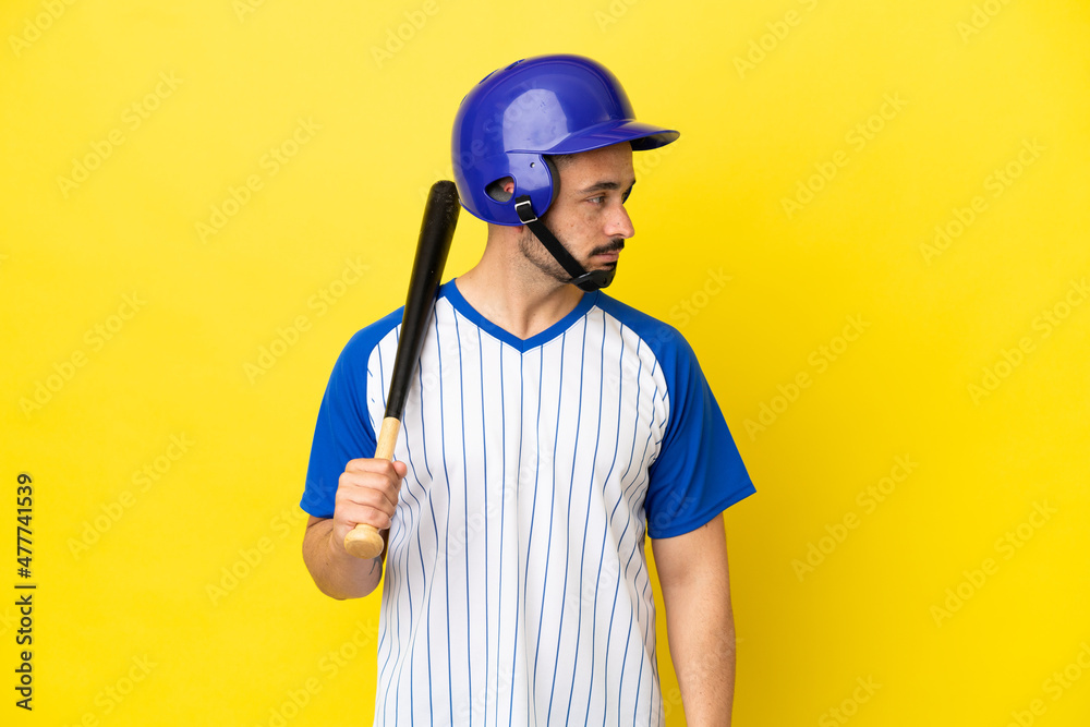 Young caucasian man playing baseball isolated on yellow background looking to the side
