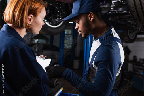 African american mechanic pointing at clipboard near colleague in car service.