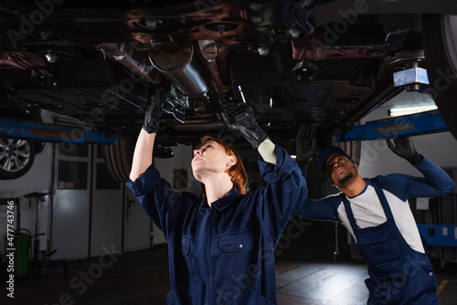 interracial technicians inspecting bottom of lifted auto in car service.
