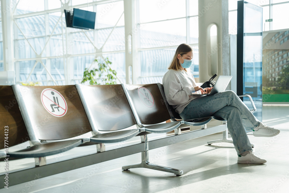 Female traveler in mask sitting in airport lobby chair with social distancing sign and working on laptop Female traveler in mask sitting in airport lobby chair with social distancing sign and working on laptop