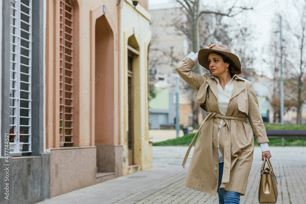 Fototapeta premium An adult woman in a beige coat and hat walking between buildings outdoors.