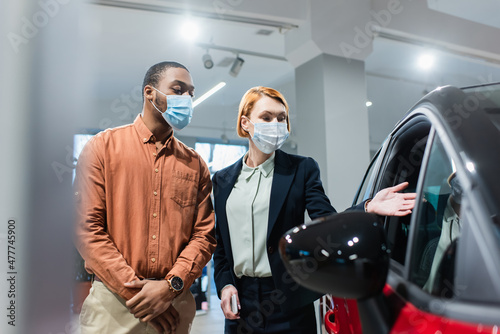 car dealer in medical mask pointing at car near african american customer.