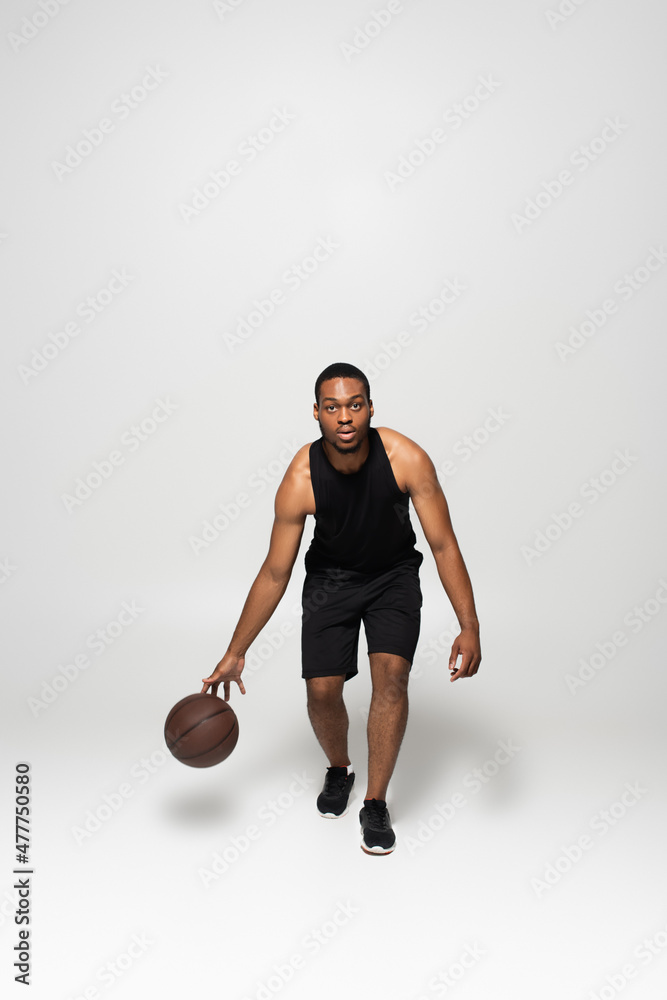 full length of muscular african american man playing basketball on grey.