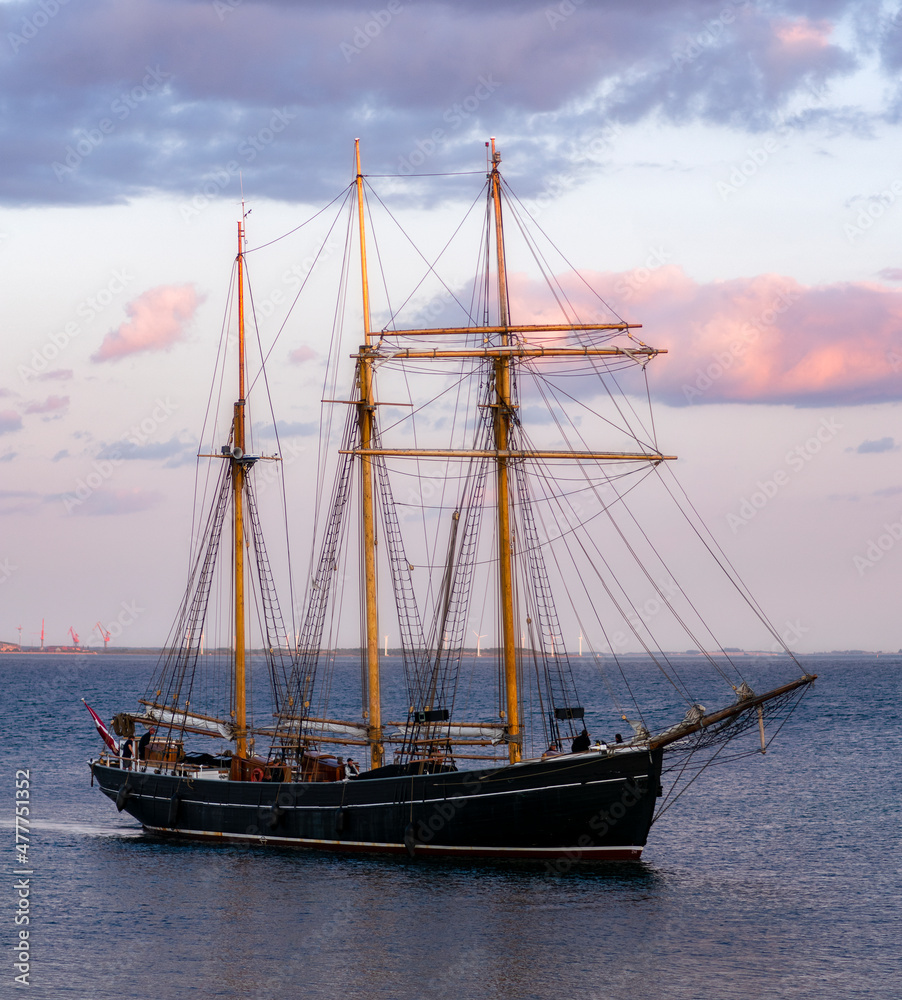 Fototapeta premium Big, old, wooden sailboat with three masts sailing in to the harbour at the little island of Ven between Sweden and Denmark in the pink light of sunset illuminating the clouds