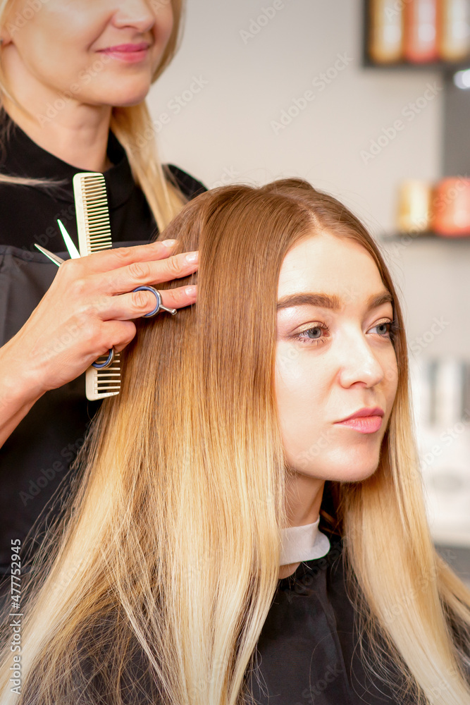 Fototapeta premium Gorgeous blonde long hair woman and her hairdresser looking and checking out hairstyle. Beautiful hairstyle of a young blonde woman after dying hair in a beauty salon, results of hair treatment