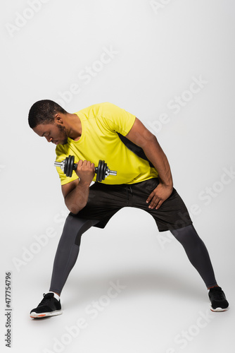 full length of african american sportsman working out with dumbbell on grey.