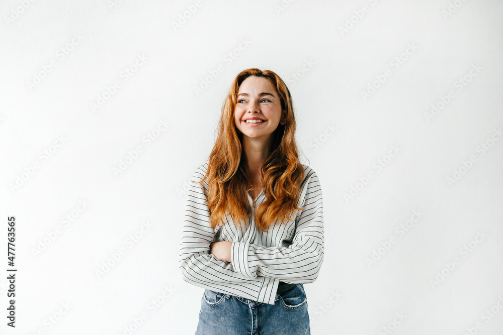 Emotive portrait of smiling redhead woman with curly hair, crossing her arms and looking to the side, standing against white background Emotive portrait of smiling redhead woman with curly hair, crossing her arms and looking to the side, standing against white background