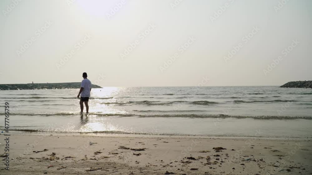 a young guy in a white T-shirt walks on the water against the backdrop of the sunset. a man admires the ocean and hits the water. city beach in the united arab emirates.