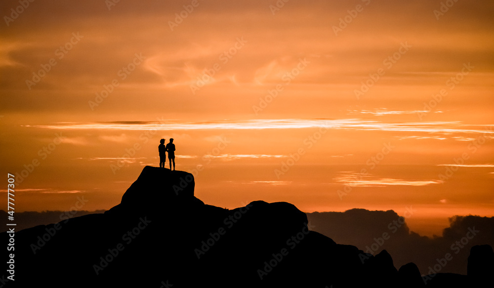 Dramatic golden sunset of two men admiring the view on vacation, standing on a large rock on a beach in Jacobsbaai, South Africa.