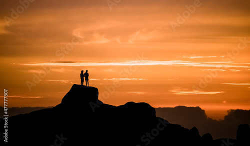 Dramatic golden sunset of two men admiring the view on vacation, standing on a large rock on a beach in Jacobsbaai, South Africa.