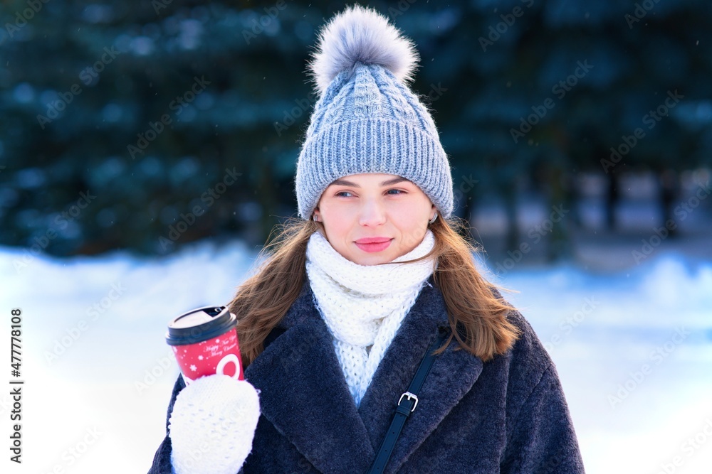 Portrait of beautiful attractive girl, happy positive joyful pretty woman in warm clothes, knitted hat and scarf at winter frosty cold snowy day is holding, drinking cup of hot tea or coffee, smiling
