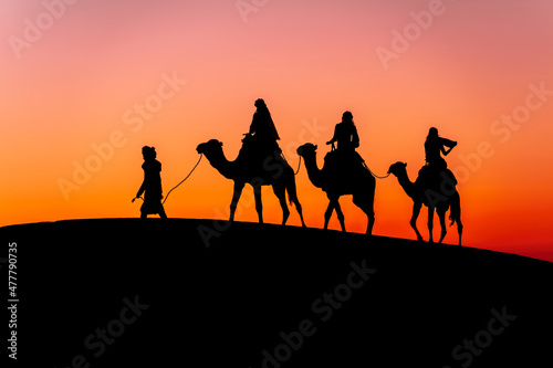 Three Riders And Their Handler Travel Through The Saharan Desert On Their Camels In Morocco