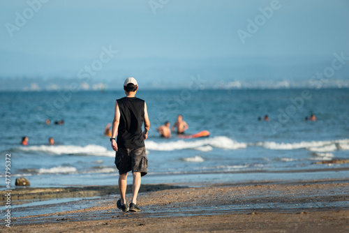 Man walking on the beach with out-of-focus people playing in the water in summer, Auckland.