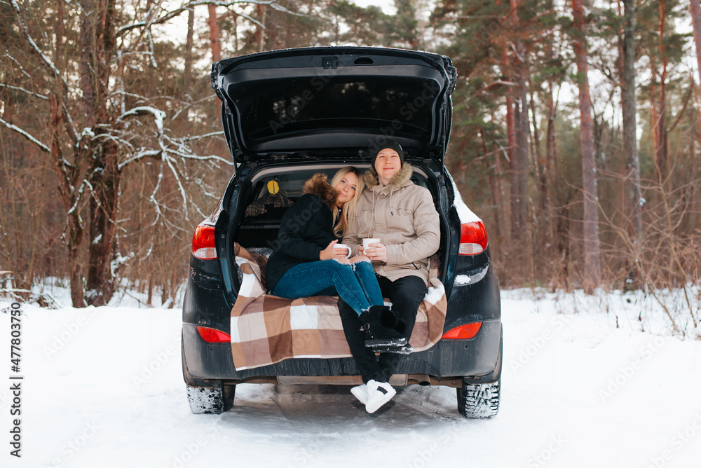 Hugging young couple of lovers having picnic in open trunk of car during journey in winter snowy forest. Happy caucasian family enjoying road trip, romantic date, outdoor recreation, valentine's day
