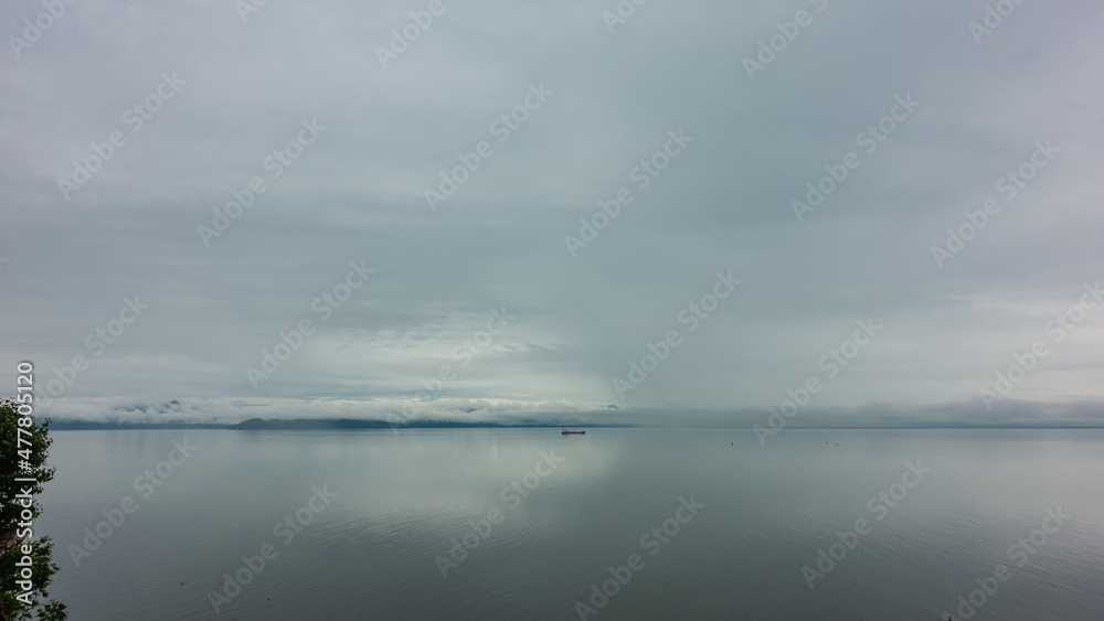 A foggy day on the Pacific Ocean. A tiny silhouette of a ship is visible on the smooth water. Cloudy sky. Reflection. Pastel shades. Copy space. Petropavlovsk-Kamchatsky