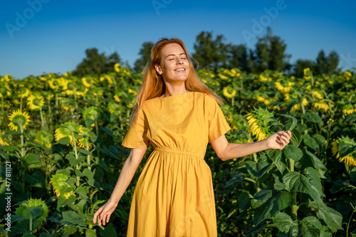 A beautiful red-haired middle-aged caucasian woman in a yellow dress enjoys the sunny weather, dancing with a closed her eyes in a sunflower field