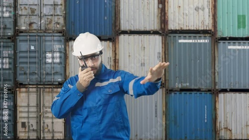 Engineer holding walkie talkie radio communication with overhead gantry crane hoist operator ready ship stacked container for import export shipping at jetty pier  
