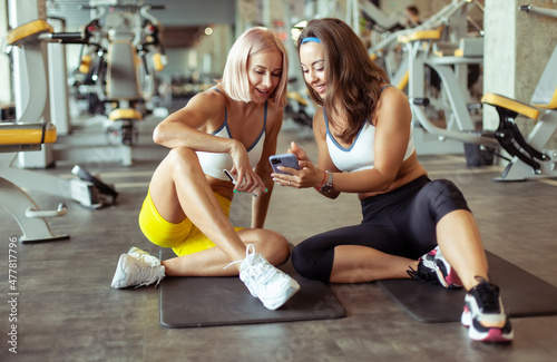 Two smiling athletic women friend looking at the smartphone screen while relaxing on the mat in the gym