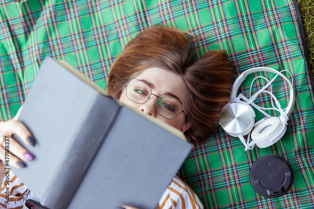 Obraz premium Young woman student reads a book while lying on a blanket in the park