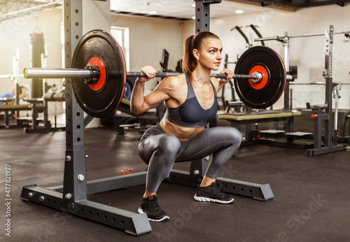 Young athletic woman practicing squats with a heavy barbell on her shoulders in the gym